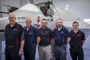 Five people in matching dark shirts with red and white trim, standing in front of a white aircraft with the number “46-003” on it. Their faces are obscured by brown rectangles for privacy.