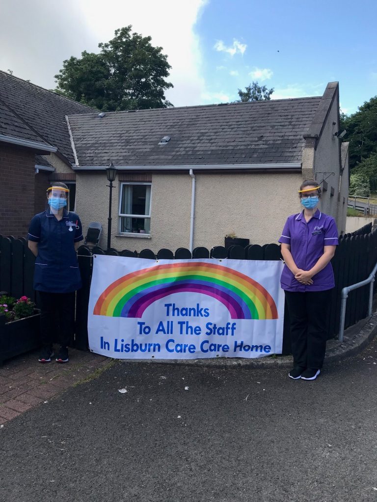 Two individuals standing beside a large banner with a rainbow and the text ‘Thanks To All The Staff In Lisburn Care Care Home,’ displayed outside a building.