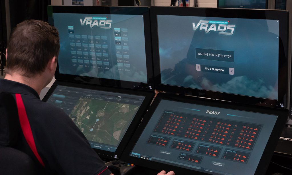 A person from behind viewing multiple computer monitors displaying a simulation program named VRADS, with one screen showing a map and another waiting for an instructor.
