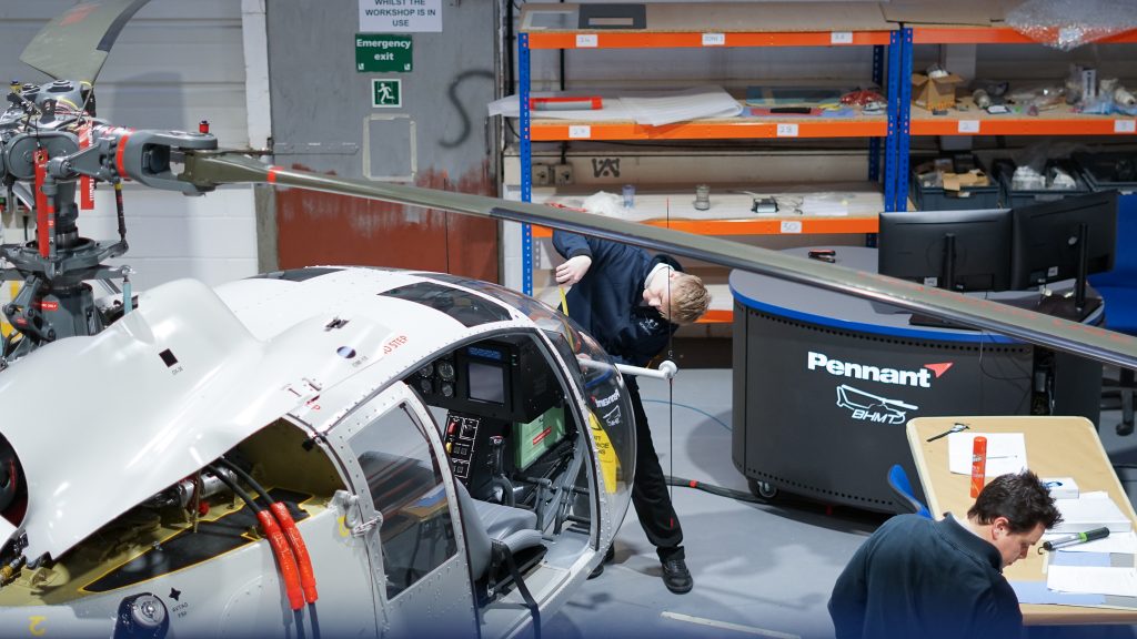 A metal training device of a helicopter in a workshop. There are people working on the training device and there are large orange shelfs in the workshop.