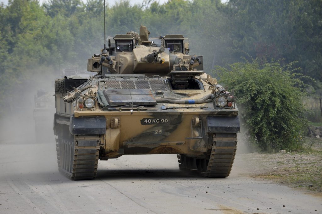 A defence army tank. There is trees and a tarmacked road