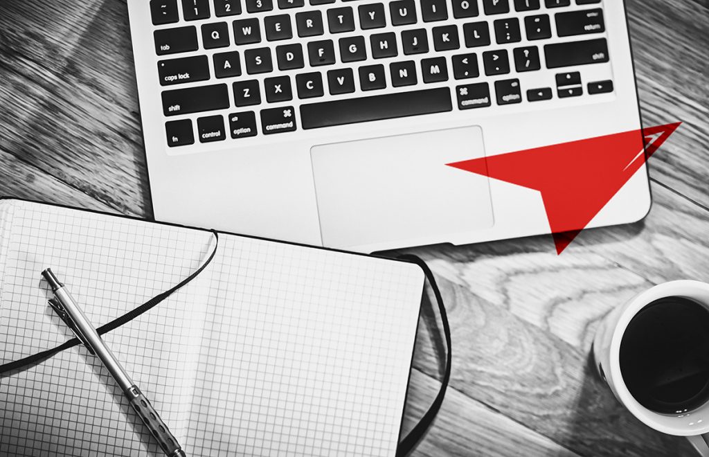 A grayscale image of a workspace with a laptop, a notepad with a pen on top, and a cup of coffee to the side. A red paper plane graphic is superimposed on the image, pointing towards the laptop’s trackpad.