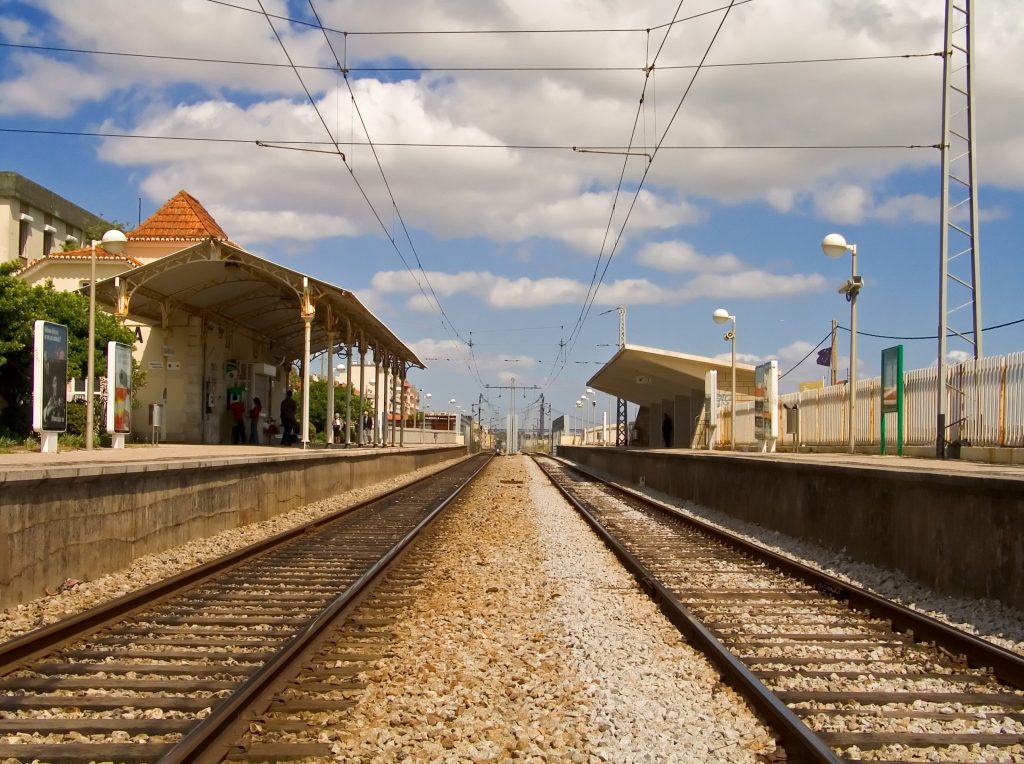A sunny railway station with two platforms, each equipped with shelters for waiting passengers. The blue sky overhead features scattered clouds. Two parallel sets of railway tracks extend into the distance, converging at a point on the horizon. Passengers are visible on the left platform, and the platform shelters have terracotta tiled roofs and white columns. Various lampposts and signs line both platforms, and a fence separates the far platform from adjacent land.