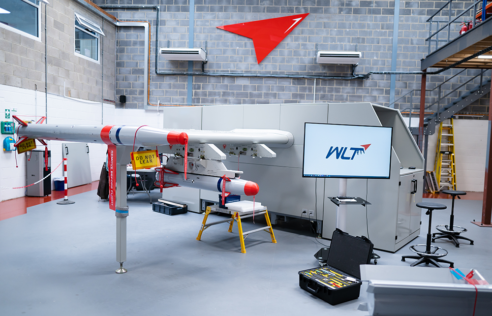 A large industrial machine with ‘WLT’ branding is the central focus in an indoor workshop setting. The machine has a long, cylindrical part extending from it, resembling a missile or rocket. There are warning signs and tools around the machine.