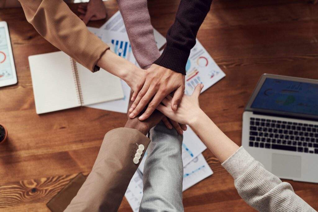 The image depicts a group of people stacking their hands in the center, suggesting unity or teamwork. The hands are of different skin tones, implying diversity. There are business-related papers with charts and graphs scattered on the wooden table around their hands. A laptop is also visible on the table, indicating a work or meeting environment. The sleeves of the individuals suggest a professional or semi-formal attire.