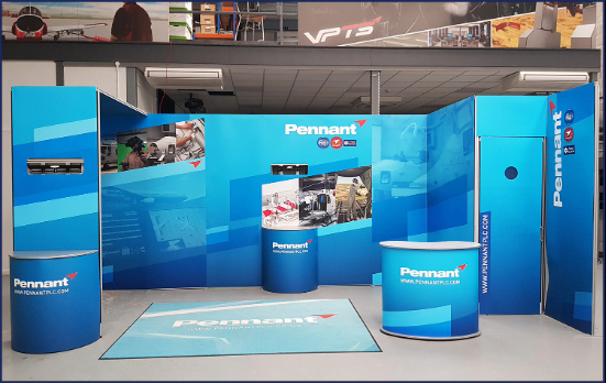 A mock-up design of an exhibitor stand in a large warehouse room. The stand in a multitude of blue colours with the word "PENNANT" on. There are various images on the panels of the stand.