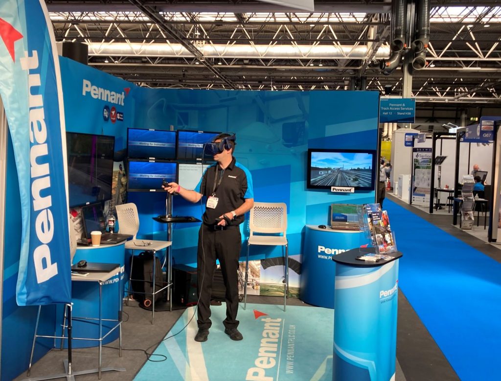 An exhibition stand set-up at an event. There display is showing a blue backdrop and there is someone in the middle with a VR headset on it.