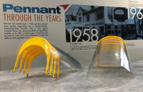 A pair of yellow and clear safety glasses lying on a reflective surface with a historical timeline of the Pennant company in the background.