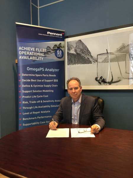 A person sat at a dark wooden desk signing pieces of paper. The person is wearing a suite and there is a pop-up banner and image on the wall behind him.