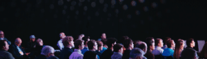 A crowd of people sat in a dark theatre conference room look in the same direction