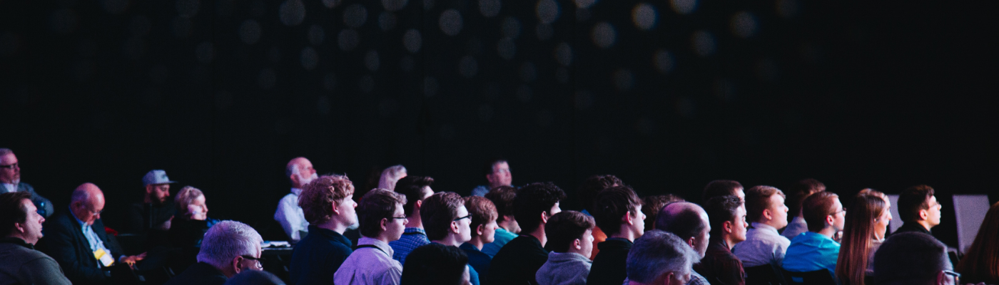 A crowd of people sat in a dark theatre conference room look in the same direction