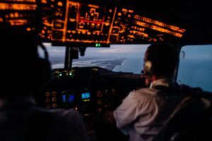 An over the shoulder image of two pilots flying an aeroplane. The cockpit is liite up and and you can see a view of a shore line