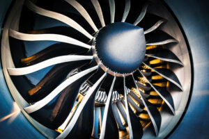 Close-up view of a jet engine with metallic fan blades arranged in a circular pattern. The center of the engine is capped with a rounded, dark-colored cover. Intricate mechanical components are partially visible behind the fan blades
