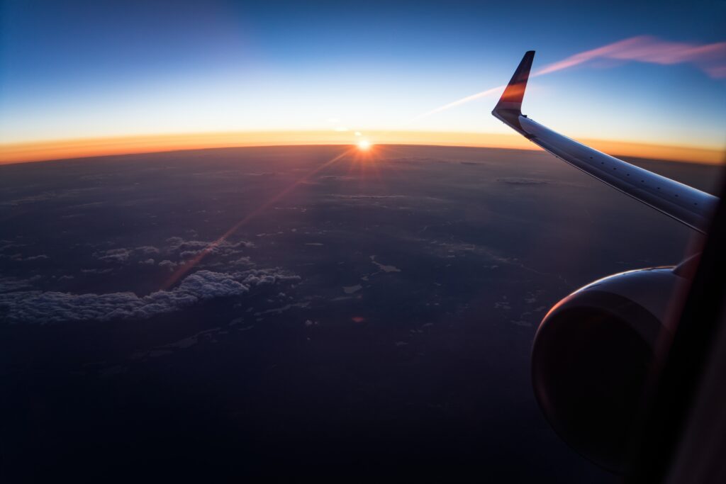 A breathtaking view from an airplane window during sunset, where the sun casts a warm glow against the darkening sky, illuminating the clouds and earth below.