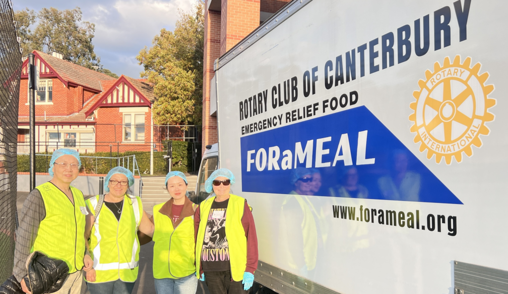 A close up shot of four smiling people wearing high-vis jackets and hair nets, stood next to a volunteering food delivery van.