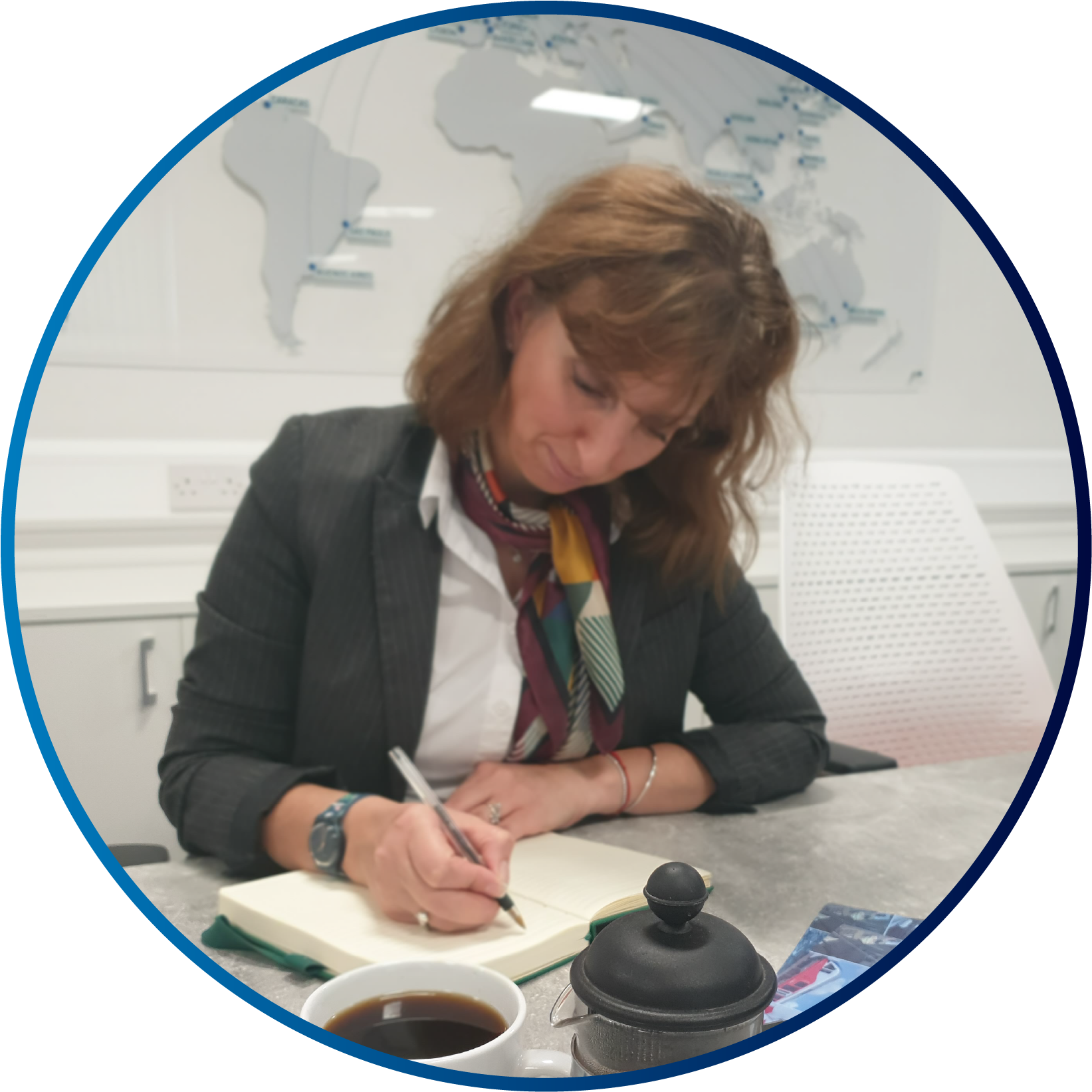 A circle image of a smartly dressed woman, sat at a desk, writing in a notepad 